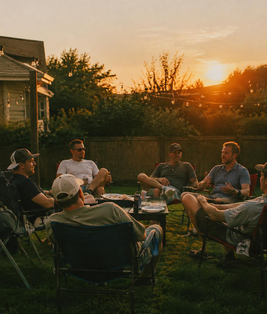 Dads in folding chairs around a low table at a backyard cookout at sunset